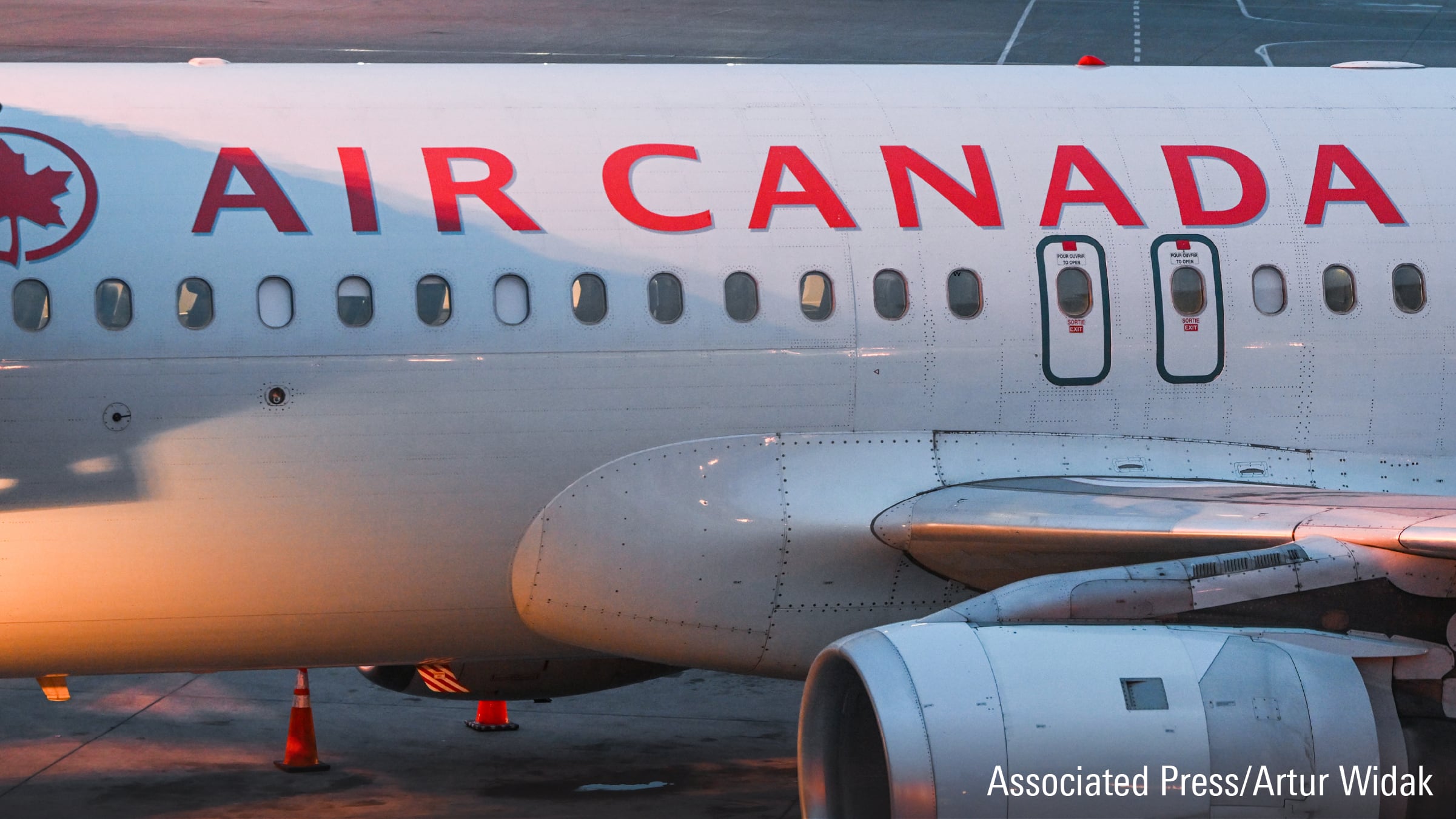 Side view of an Air Canada airplane on the tarmac, showing the logo, windows, exit doors, and part of the engine in soft sunlight. Orange cones sit below.